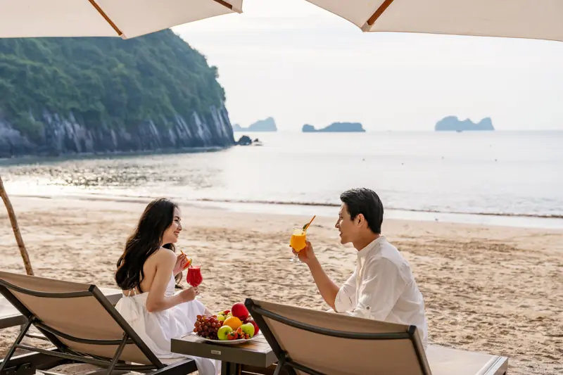 Hai người ngồi ghế nằm dưới ô trên bãi Cát Cò, phía xa là các đảo đá