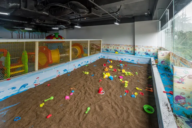 Hố cát trong nhà ở Kids Club với xẻng, xô đồ chơi nhiều màu, phía sau là khu cầu trượt và ống chui