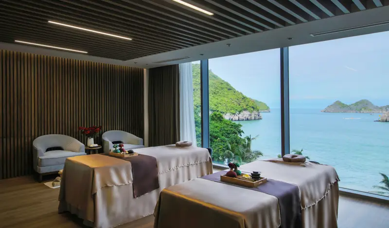 Phòng spa đôi với hai giường trải khăn, ghế bành và vách kính lớn nhìn ra biển xanh và núi đá