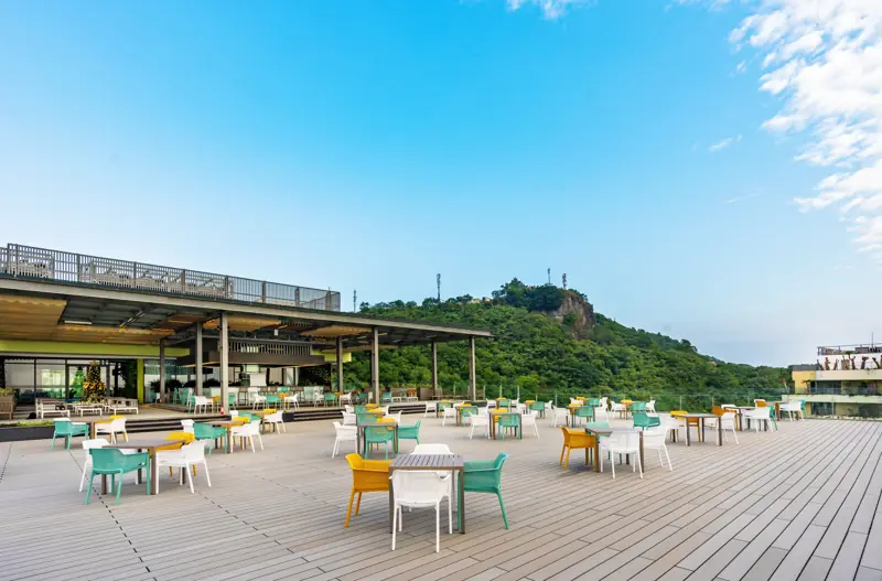 Sân thượng Lan Hạ Sky Bar sàn gỗ với bàn ghế xanh, vàng, mái che kính và đồi cây xanh phía sau dưới trời xanh