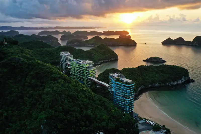 Hoàng hôn trên vịnh Lan Hạ nhìn từ trên cao: các đảo đá vôi nhấp nhô, các tòa nhà Flamingo giữa rừng xanh và bãi cát cong bên dưới