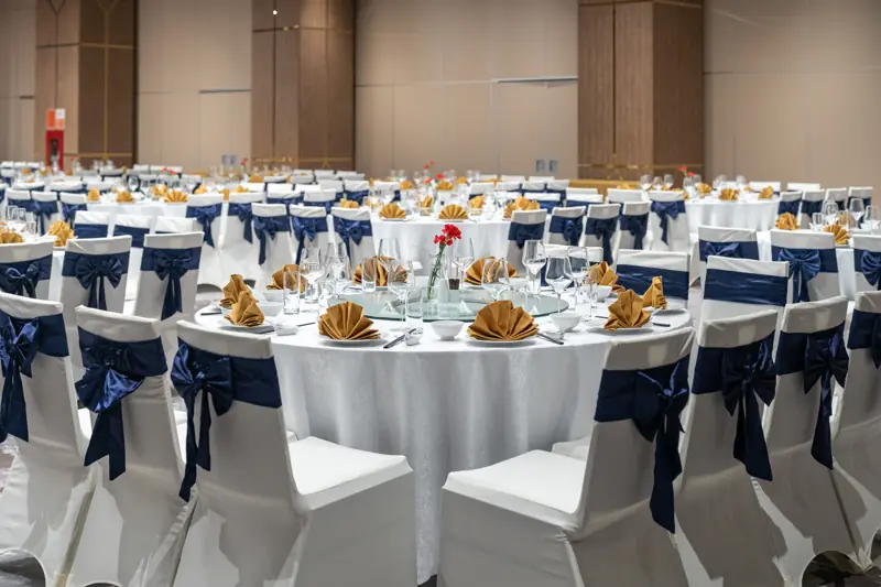 Bàn tiệc tròn trong Grand Opera phủ khăn trắng, ly pha lê, khăn ăn gấp quạt vàng, ghế thắt nơ xanh navy