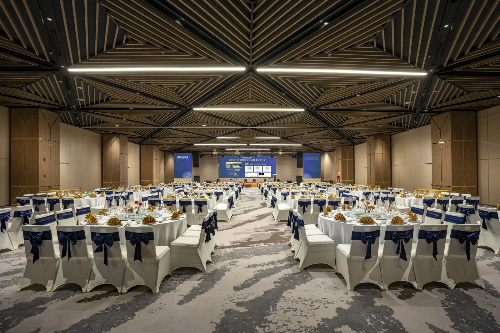 Hội trường Grand Opera ở Flamingo Cát Bà bày hàng trăm bàn tiệc tròn, ghế bọc trắng thắt nơ xanh navy, trần gỗ xếp hình kim cương