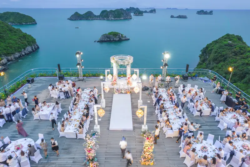 Sân thượng Lan Hạ Sky Bar nhìn từ trên cao: các dãy bàn tiệc kín khách, lối đi trắng giữa, phía sau là vịnh Lan Hạ và đảo đá