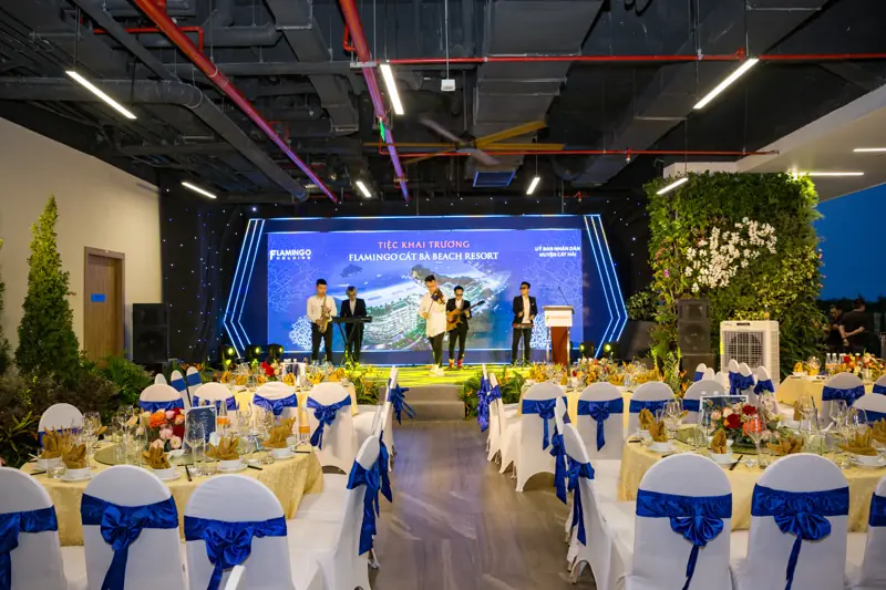 Tiệc khai trương ở Sky Garden: ban nhạc trên sân khấu trước màn LED, bàn tiệc ghế trắng nơ xanh, tường cây xanh bên phải