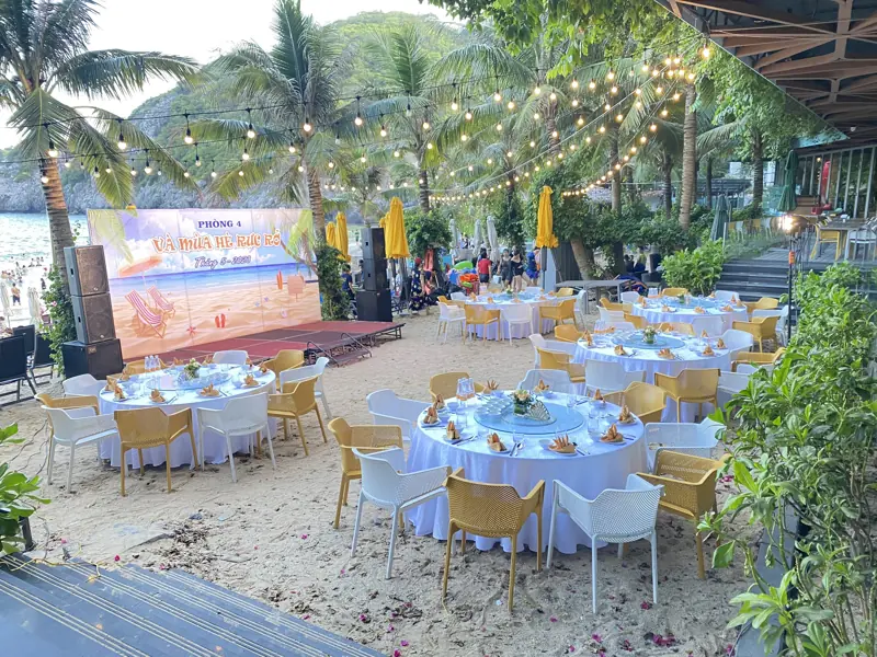 Tiệc trên bãi cát dưới hàng dừa ở Flamingo Cát Bà: bàn tròn khăn trắng, ghế vàng, dây đèn bóng tròn và sân khấu nhỏ sát biển