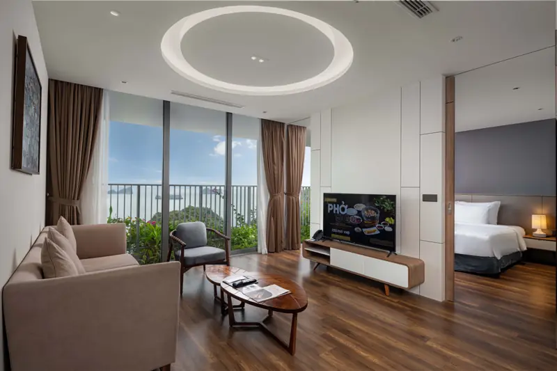 Phòng khách căn Deluxe Residence 1 phòng ngủ: sofa, ti vi, cửa kính ra ban công nhìn biển, phòng ngủ phía sau