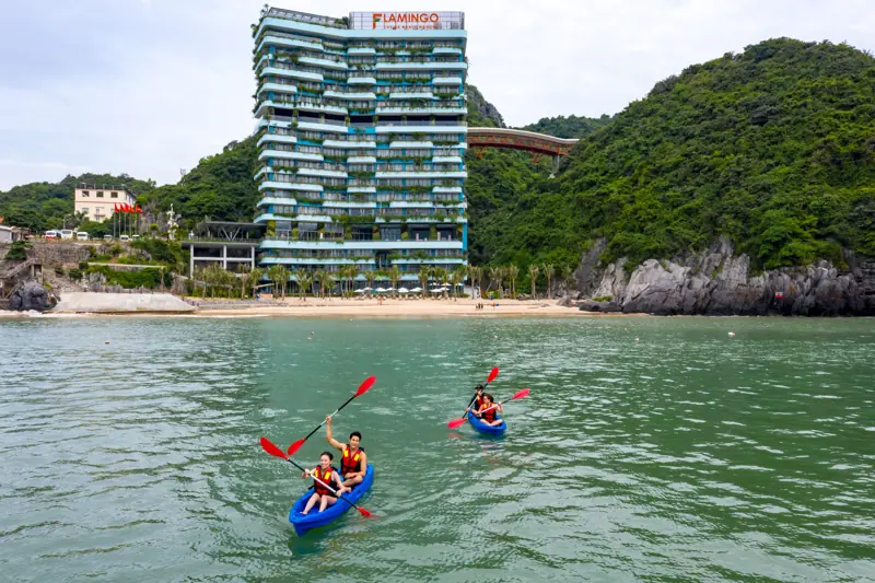Hai thuyền kayak xanh chèo trên mặt nước xanh ngọc trước bãi Cát Cò, phía sau là tòa nhà phủ cây và núi đá