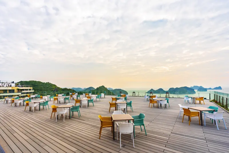 Sân thượng Lan Hạ Sky Bar bày bàn ghế ngoài trời, phía xa là vịnh lúc chiều
