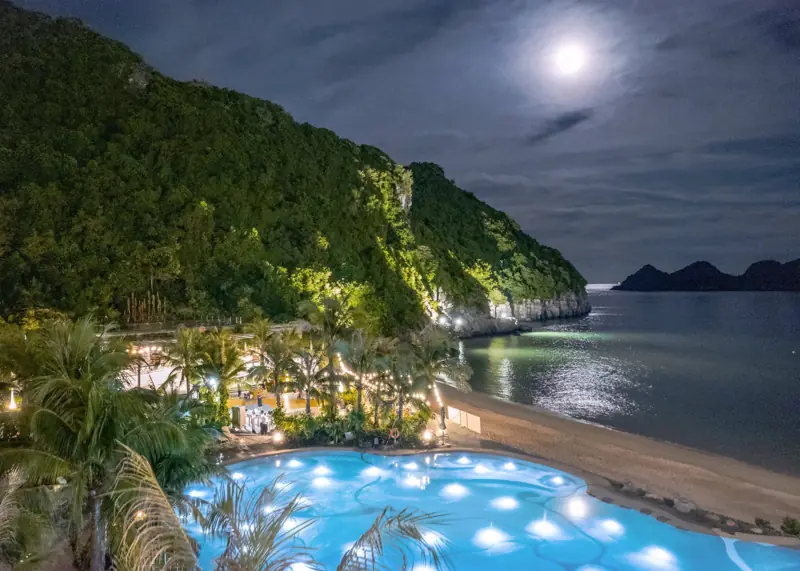 Bể bơi Charm Pool lên đèn xanh ban đêm dưới chân vách núi, bên cạnh bãi Cát Cò và vầng trăng trên biển
