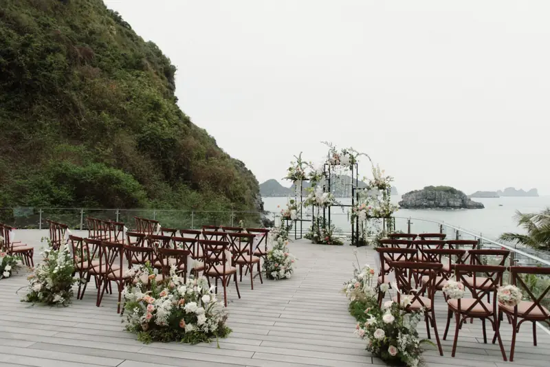 Sàn gỗ ngoài trời bày ghế gỗ và hoa cho lễ cưới, cổng hoa nhìn ra biển và đảo đá dưới chân vách núi