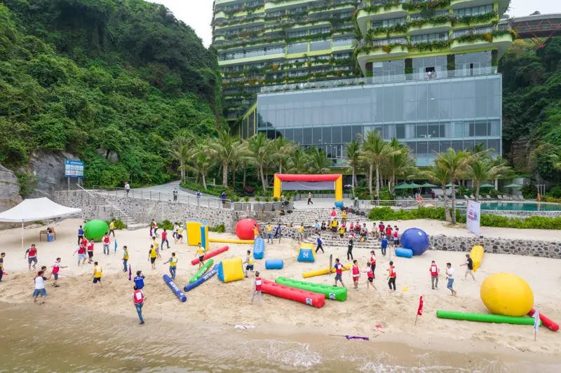 Đoàn khách chơi trò vận động với dụng cụ hơi nhiều màu trên bãi Cát Cò, phía sau là tòa khách sạn phủ cây xanh